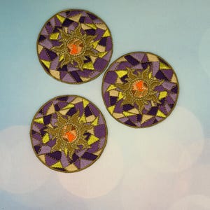 May include: Three embroidered circular coasters with a sunburst design. The coasters feature a central orange and brown sun surrounded by a gold outline and a pattern of purple, yellow, and beige geometric shapes. The coasters are approximately 4 inches in diameter.