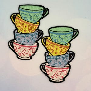 May include: Two embroidered patches showing stacks of teacups. Each stack has five cups in green, yellow, blue, and red, with black outlines. The cups are decorated with various patterns.