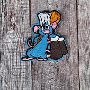Può includere: Una toppa ricamata con un ratto dei cartoni animati che indossa un cappello da cuoco e porta una borsa marrone.