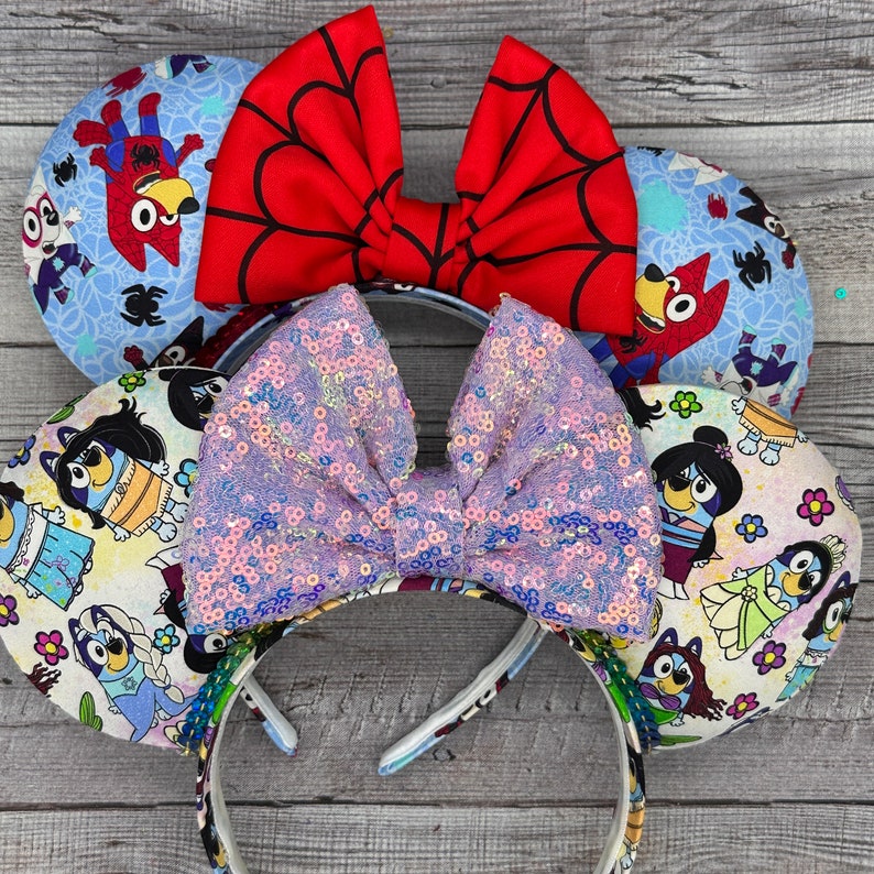 Może przedstawiać: Para uszu Minnie Mouse w kolorze fioletowym i białym z błyszczącymi elementami, ozdobiona czerwoną, białą i niebieską kokardką oraz niebieską, białą i czerwoną kokardką w kształcie pajęczyny. Uszy są ozdobione postaciami z kresk&oacute;wki Bluey.