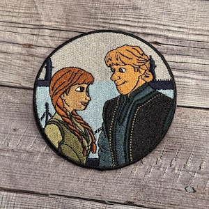 May include: Embroidered patch featuring a cartoon image of a girl with red hair and a boy with blonde hair. The girl is wearing a green dress and the boy is wearing a dark blue jacket. The background is a light blue sky with white clouds.