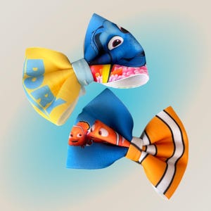 Peut inclure: Trois nœuds papillon en tissu colorés avec des personnages du film "Le Monde de Nemo". Un nœud papillon est jaune avec le mot "DORY" imprimé dessus. Les deux autres sont bleus et oranges, avec des images de Nemo et Dory.