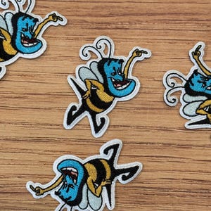 May include: Embroidered patches featuring a cartoon bee with a blue head, yellow body, and black stripes. The bee is smiling and has its arm raised. The patches have a white border and are on a wooden surface.