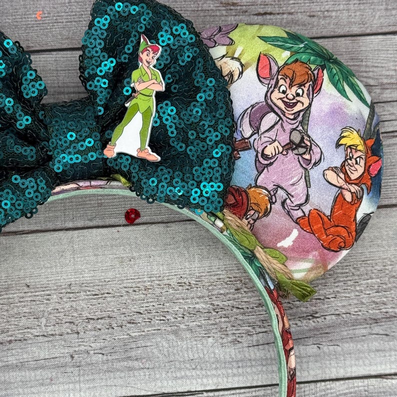 May include: A teal sequined bow with a Peter Pan character pin is attached to a headband. The headband features a colorful print of Peter Pan characters. The bow is embellished with sequins.