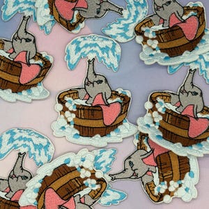 May include: Multiple embroidered patches featuring a grey elephant with large pink ears sitting in a brown wooden tub. The elephant is spraying a blue and white water fountain from its trunk, surrounded by white and light blue soap bubbles.