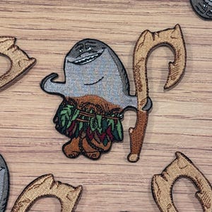 May include: Embroidered patch featuring a smiling, gray cartoon character with a brown and green skirt, holding a brown hook-shaped weapon. The patch is on a wooden surface, with other patches visible.