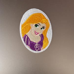 Peut inclure: Un écusson brodé ovale blanc représentant un dessin animé de Raiponce du film d'animation Disney Tangled. Elle a de longs cheveux blonds, une robe violette et un sourire doux.