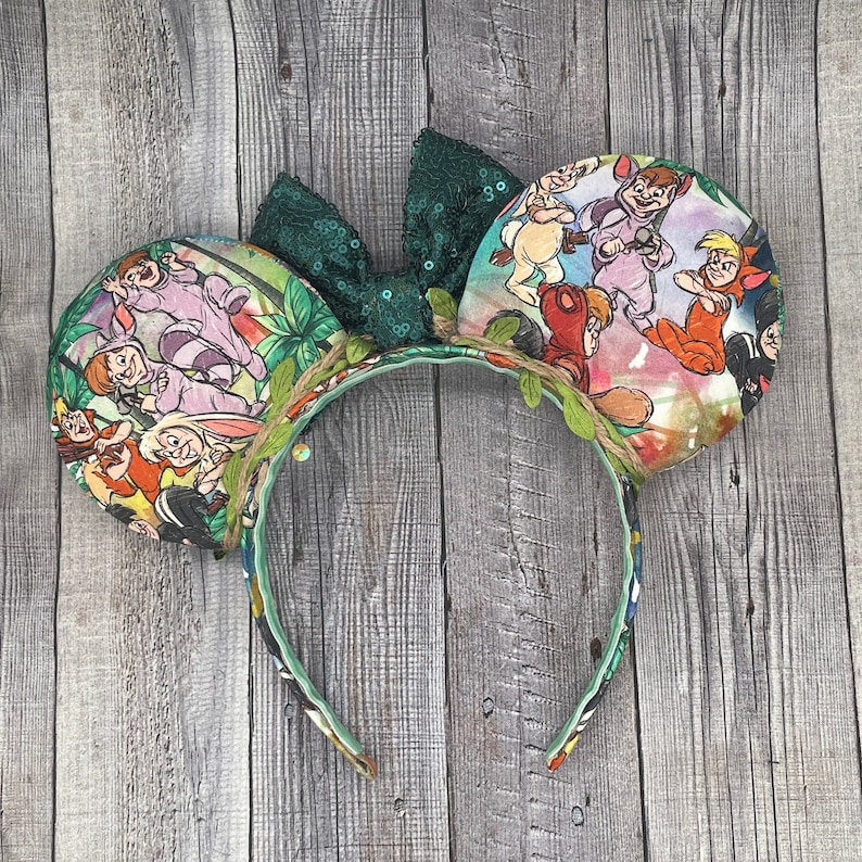 May include: A pair of mouse ears featuring a Peter Pan theme. The ears are decorated with colorful images of Peter Pan characters. A green sequined bow sits between the ears, and the headband is adorned with faux vines and leaves.