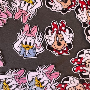 Può includere: Toppe ricamate con Paperina e Minnie. Paperina indossa un top blu e un fiocco rosa. Minnie indossa un vestito rosso a pois bianchi e un fiocco abbinato. Entrambi i personaggi fanno il segno della pace.