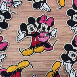 May include: Embroidered patches featuring Mickey Mouse and Minnie Mouse in a loving embrace. Mickey wears a red shirt and yellow shoes, while Minnie dons a pink dress and bow. The patches have a cream-colored border and are scattered on a wooden surface.