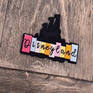 May include: A black embroidered patch featuring a silhouette of Sleeping Beauty Castle and the text "Disneyland" in rainbow colours.