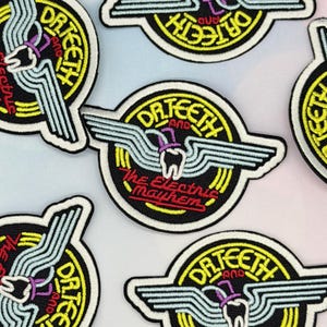 May include: Circular embroidered patches featuring the text Dr. Teeth and The Electric Mayhem. The design includes a central white tooth wearing a purple top hat, flanked by light blue wings, set against a black background with yellow rings.