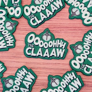 May include: Green and white embroidered patches with the text "Oooooohhh The Claaaw" and a claw graphic. The patches are scattered on a light brown wooden surface. The patches have a green background with white lettering and a dark green border.