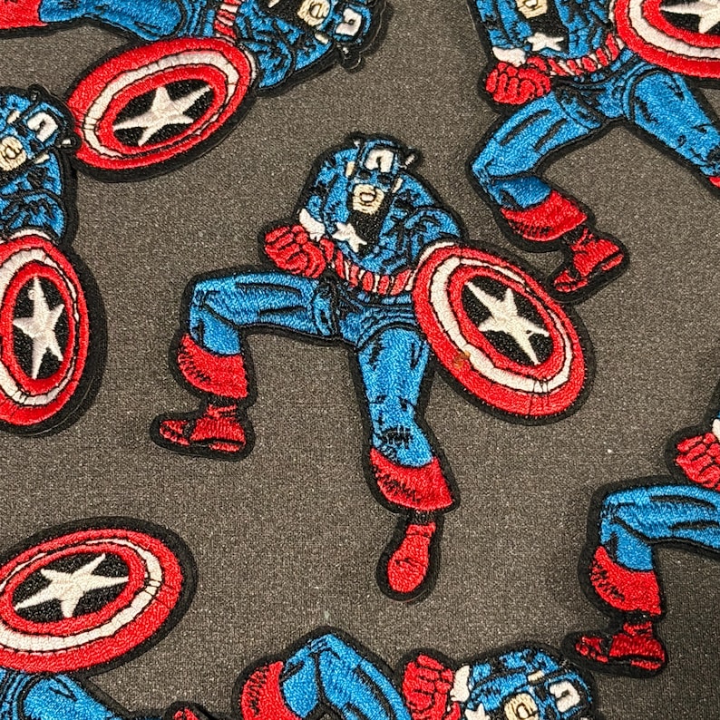 May include: Embroidered patches of Captain America in action, holding his shield. The patches are predominantly blue, red, and white, with intricate stitching. The shield features a white star on a red backdrop. The patches are set against a dark grey background.