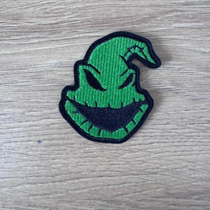 May include: Green embroidered patch of the character Oogie Boogie from the movie The Nightmare Before Christmas. The patch features a green face with a black mouth and eyes.