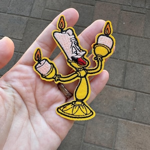 May include: Yellow embroidered iron-on patch of a cartoon candlestick character with a smiling face and three candles. The character is wearing a yellow suit and has a red tongue sticking out.