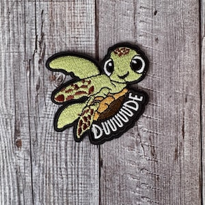 Pode incluir: Um remendo bordado com uma tartaruga marinha de desenho animado em verde e marrom com a palavra "DUDE" escrita em uma fonte ondulada.