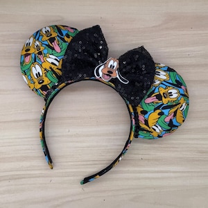 Può includere: Un fiocco nero con paillettes e il personaggio di Pluto adornano una fascia per capelli con orecchie di Topolino. Le orecchie presentano un motivo colorato del volto di Pluto su uno sfondo blu, verde e giallo. La fascia è nera con un motivo colorato.
