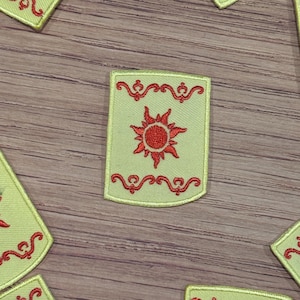 Op de afbeelding: Een groene geborduurde patch met een rood zonontwerp. De zon heeft een rood midden en acht rode stralen. De patch heeft een rode rand met een decoratief ontwerp.