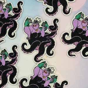 Op de afbeelding: Meerdere geborduurde patches met een personage met wit haar, een paarse huid en een zwart onderlichaam van een octopus. Het personage heeft groene aalachtige wezens op elke schouder, een gouden schelpenketting en een brede grijns met rode lippen.