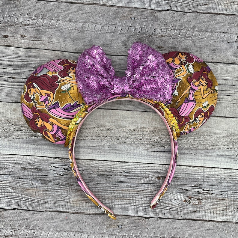 K&ouml;nnte beinhalten: Mausohren-Stirnband mit einer lila Pailletten-Schleife und Ohrendetails mit einer Zeichentrickfigur in einem rosa Kleid. Das Stirnband ist rosa mit goldenen Akzenten.