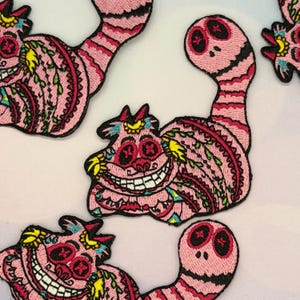 May include: Pink embroidered patches featuring the Cheshire Cat from Alice in Wonderland. The patches have a detailed design with a smiling face, stripes, and floral patterns. The patches are outlined in black.