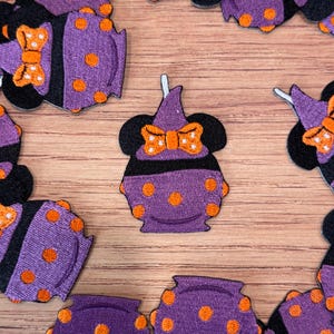 May include: Embroidered purple cauldron patches with orange polka dots and a black witch hat design. Each patch features a bow and mouse ears. The patches are arranged on a wooden surface.
