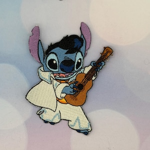May include: Embroidered patch featuring the character Stitch dressed as Elvis Presley, playing a brown guitar. Stitch is blue with large eyes, wearing a white jumpsuit and a black pompadour hairstyle. The patch is suitable for clothing or accessories.