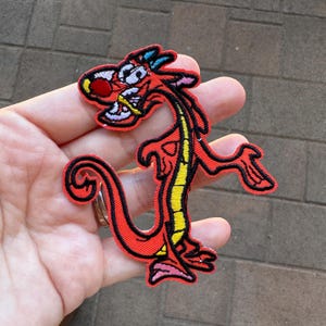 May include: Embroidered patch featuring a cartoon dragon in red, yellow, and black. The dragon has a red body, yellow spine, and black outline. The patch is held in a hand against a textured background.