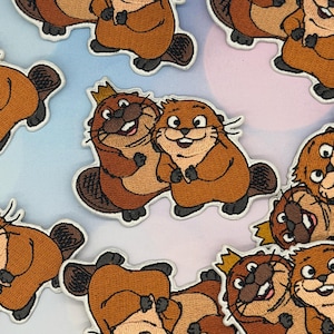 May include: Embroidered patches of cartoon beavers in brown, tan, and black. Some beavers wear crowns. The patches have a white border and are arranged on a light blue and pink background.