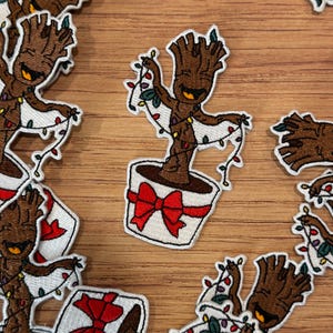 May include: Embroidered patch featuring a cartoon tree-like character decorated with Christmas lights, planted in a white pot with a red bow. The character has a smiling face and is surrounded by other patches.