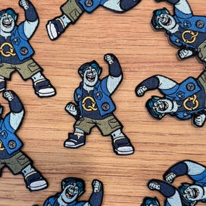 May include: Embroidered patches featuring a cartoon character in a blue jacket, khaki pants, and black shoes. The patches have a circular emblem with a "Q" on the jacket. The patches are scattered on a wooden surface.