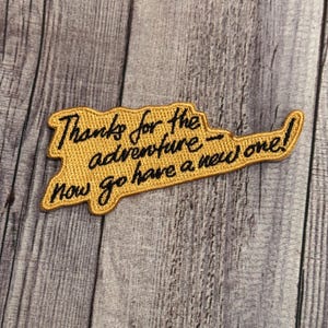 May include: A gold embroidered patch with black stitching that says "Thanks for the adventure - now go have a new one!"