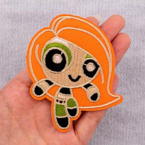 May include: An embroidered iron-on patch featuring a cartoon girl with orange hair, green eyes, and a black and green outfit.