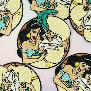 Peut inclure: Écussons brodés représentant la princesse Jasmine et un personnage avec une barbe blanche et un turban. Les écussons ont un fond crème avec des contours noirs et sont de forme circulaire. Les personnages portent des vêtements bleus et dorés.
