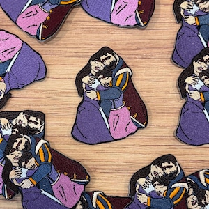 May include: Embroidered patches featuring a cartoon depiction of a family embracing. The patches show a man, woman, and child in period clothing, with purple, pink, and blue hues. The patches are scattered on a wooden surface.
