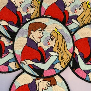 May include: Embroidered patches featuring a romantic scene of a prince and princess embracing. The patches are circular, with a light blue, pink, and white cloud-like background. The prince wears a red jacket and the princess has blonde hair.