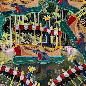 May include: Embroidered patches featuring a riverboat with a red and white striped awning, a ship's wheel, and a hippo with an open mouth. The patches are set against a background of water and trees.