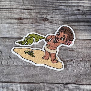 May include: An embroidered patch featuring a cartoon character holding a green leaf. The character is brown-skinned and has dark hair. The character is wearing a brown lei and a white skirt. The patch is set on a white background with a brown sand-like texture. A small green turtle is also embroidered on the patch.