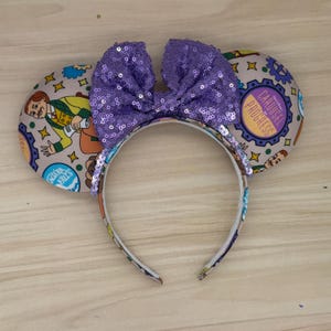 May include: A headband featuring two circular ears with a beige background patterned with cartoon characters, gears, and the text Carousel of Progress and Niagara Falls. A large, bright purple sequined bow is centred between the ears.