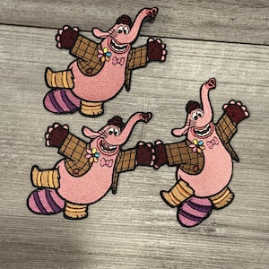 Puede incluir: Tres parches bordados con un personaje de elefante rosa con sombrero, chaqueta y guantes marrones. El elefante tiene una cara sonriente, una pajarita y una flor. Los parches están sobre una superficie de madera gris.