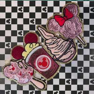 Könnte beinhalten: Vier gestickte Bügelbilder mit Disney-Charakteren und Lebensmitteln. Die Patches sind rosa, rot, weiß und gelb mit goldfarbenem Glitzerbesatz. Die Patches beinhalten eine rosa Zuckerwatte mit einer roten Schleife, einen rosa Eisbecher, ein rosa Mickey Mouse Eis am Stiel und einen roten Mickey Mouse Popcorn-Eimer.