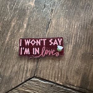 May include: A burgundy embroidered patch with pink thread that says "I won't say I'm in love" with a white flower with green leaves.