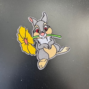 May include: Embroidered iron-on patch featuring a gray rabbit holding a yellow flower in its mouth. The rabbit has red eyes and a pink nose.