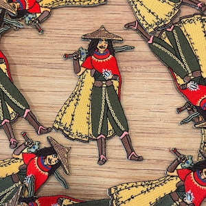 Könnte beinhalten: Gestickter Aufnäher mit einer Kriegerfigur mit Strohhut, rotem Oberteil und gelbem Umhang, die ein Schwert hält. Das Design umfasst grüne Hosen und braune Stiefel. Mehrere Aufnäher liegen auf einer Holzoberfläche.