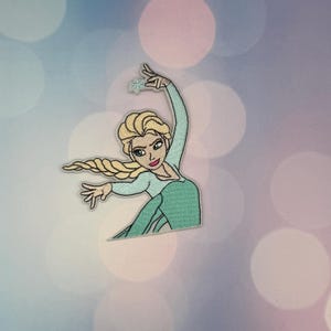 May include: An embroidered patch featuring a character with long blonde hair in a braid, wearing a light blue dress. The character has one arm raised with a small light blue snowflake icon above the hand and the other arm extended outward.