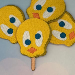 May include: Yellow embroidered patches featuring the cartoon character Tweety Bird. Each patch has a yellow face with white eyes, blue pupils, and an orange beak. One patch is shaped like a popsicle with a brown stick.