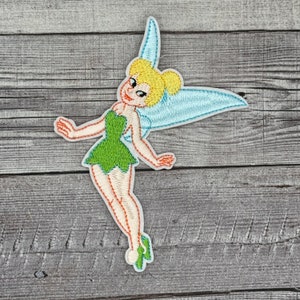 Może przedstawiać: Naszywka haftowana do prasowania z Tinkerbell, wróżką o blond włosach i zielonej sukience, z niebieskimi skrzydłami.
