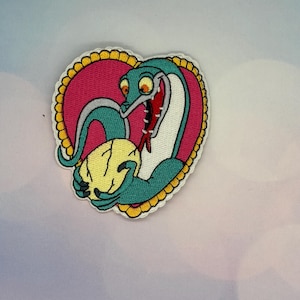 May include: Embroidered patch featuring a cartoon snake with teal and white markings, holding a yellow object. The snake is set against a pink heart-shaped background with a gold border. The snake has a red mouth and orange eyes.