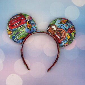 May include: A pair of mouse ears featuring a colorful Cars movie print. The ears are round and padded, with a red headband. The fabric displays Lightning McQueen and other characters, with the number 95 prominently featured.